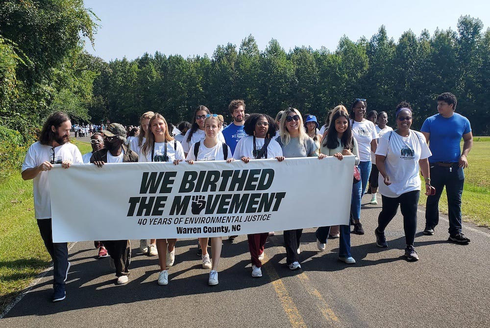 A large group, diverse in age, gender and race, march down a tree-lined road holding a sign reading "We Birthed the Movement: 40 Years of Environmental Justice."